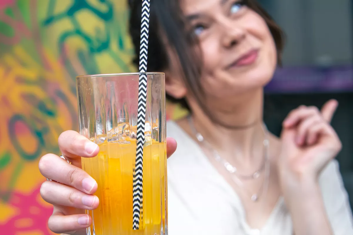 Close-up em foco de um copo de vidro com bebida alaranjada refrescante, gelo e um canudo listrado em preto e branco. Ao fundo, uma mulher sorridente e desfocada em um ambiente com grafites coloridos, reforçando o clima de festa e consumo de bebidas