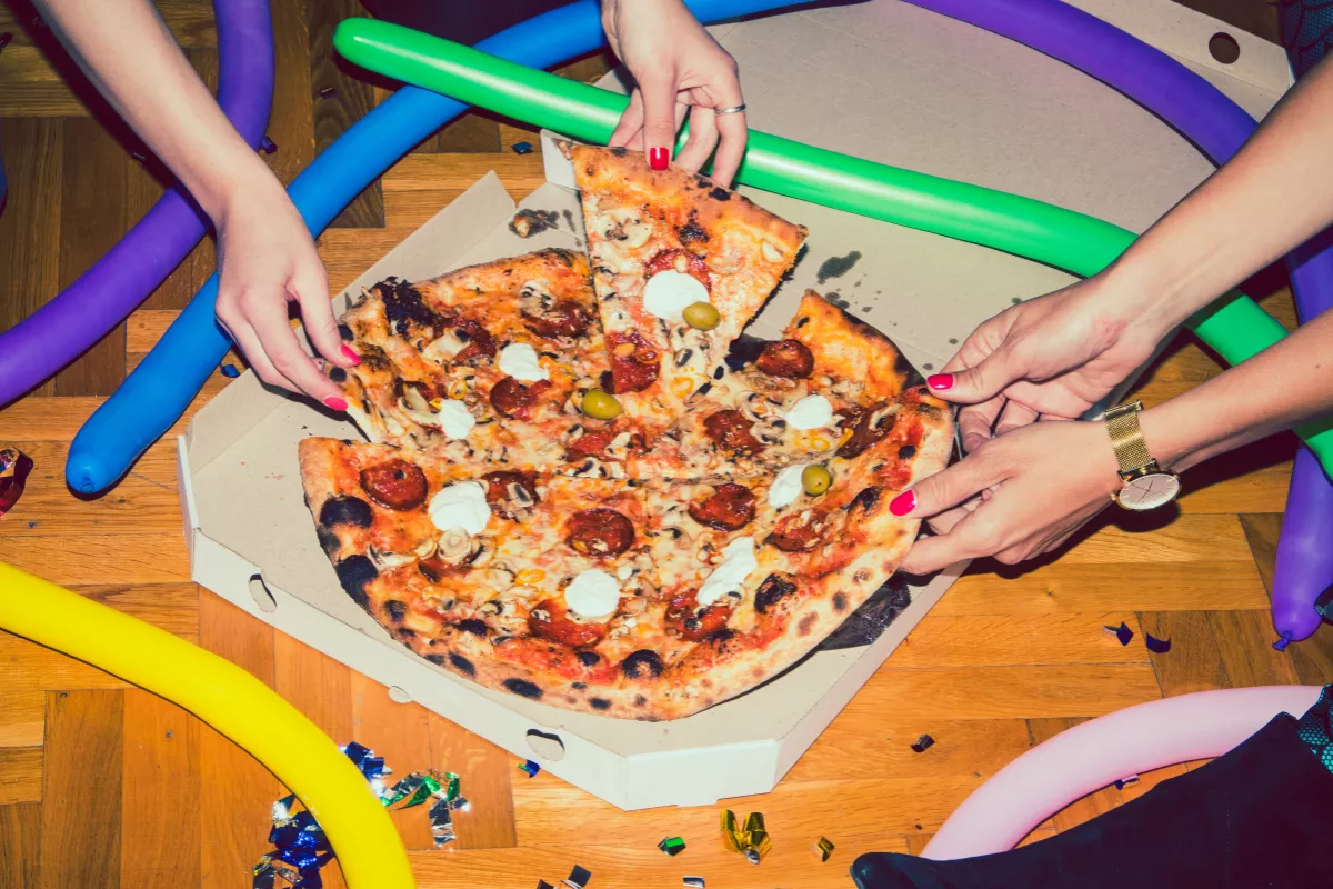 Várias mãos pegam fatias de uma pizza de calabresa e cogumelos dentro de uma caixa de papelão no chão. O ambiente está decorado com confetes, serpentinas e balões coloridos em formato de bastão, representando uma festa em casa ou 'esquenta' de Carnava