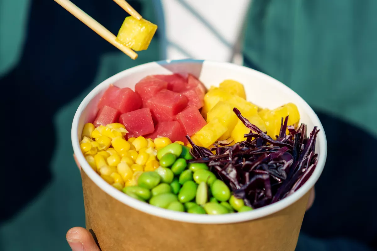 Close de um poke customizável sendo montado com hashis, contendo cubos de atum, milho, edamame e repolho roxo. Demonstração de praticidade e personalização de pedidos em restaurantes modernos.