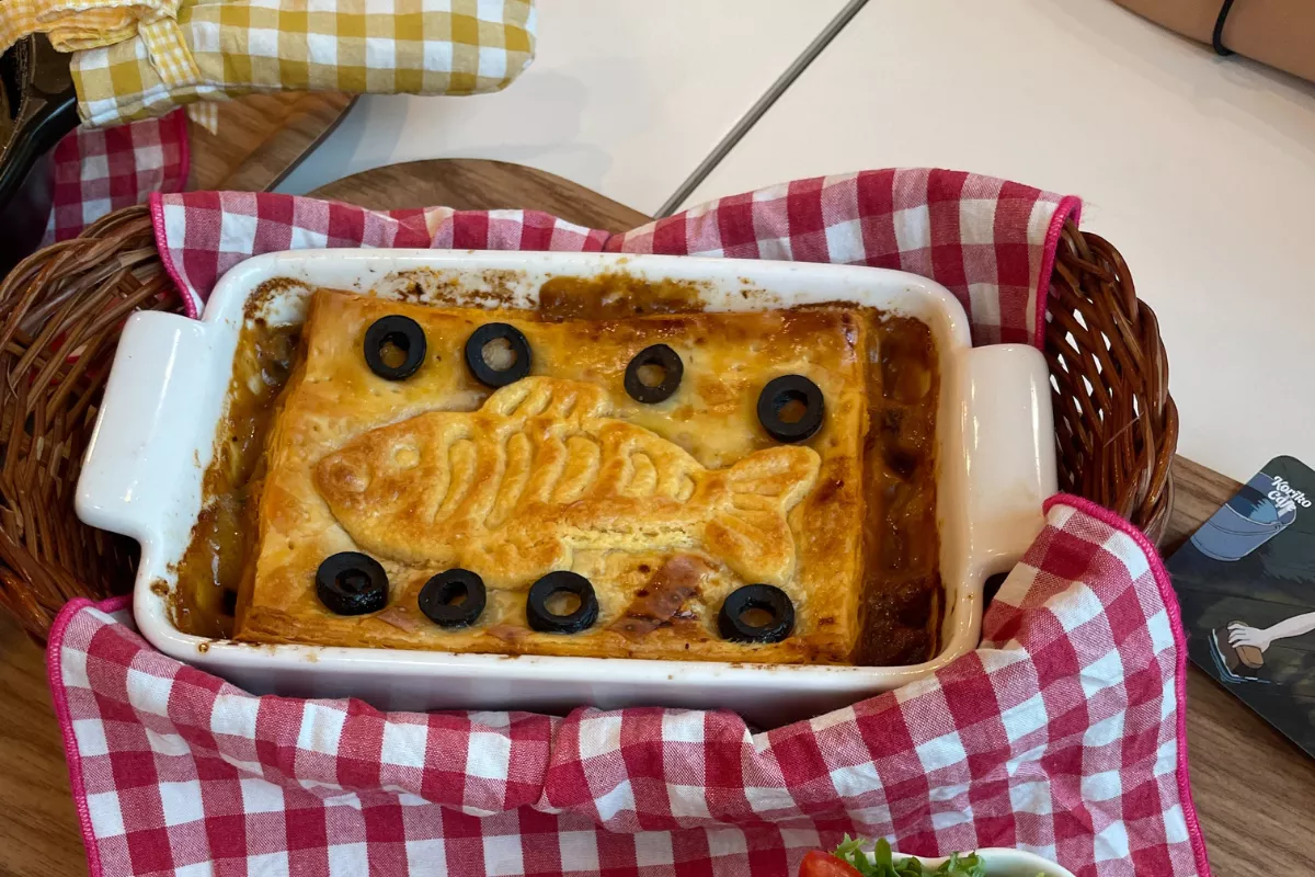 Torta salgada assada, decorada com massa em formato de peixe e azeitonas pretas, servida em uma travessa branca sobre um pano xadrez vermelho. Uma excelente opção de prato econômico e rentável para o cardápio de Quaresma em restaurantes.