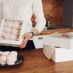 Mulher segurando uma caixa branca com visor transparente cheia de doces e suspiros, com outras caixas empilhadas sobre uma mesa de madeira, representando a entrega programada de clubes de assinatura.