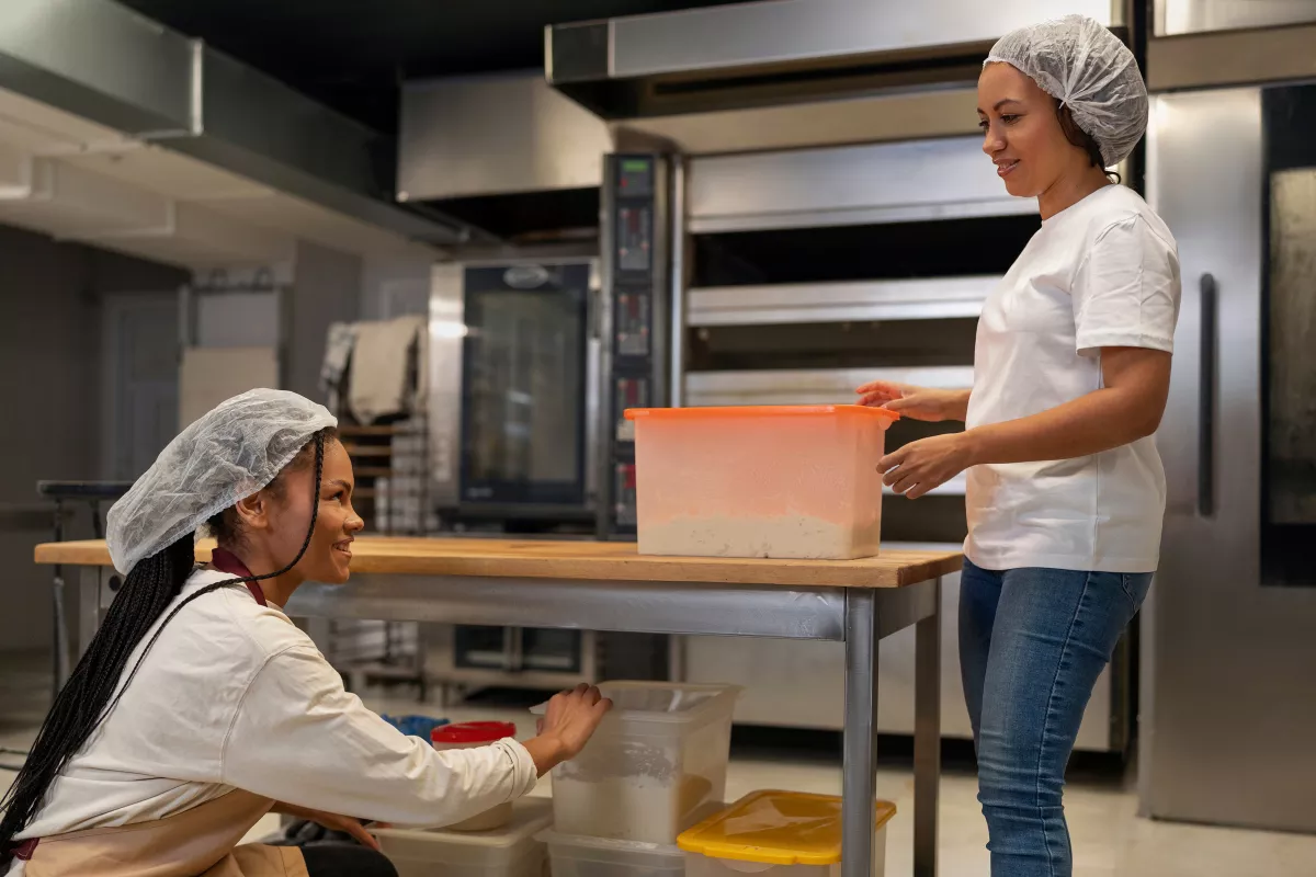 Duas funcionárias usando touca e uniforme branco organizando caixas de ingredientes de plástico transparente em uma bancada de inox dentro de uma cozinha industrial. O controle de estoque eficiente evita o desperdício de insumos no restaurante.