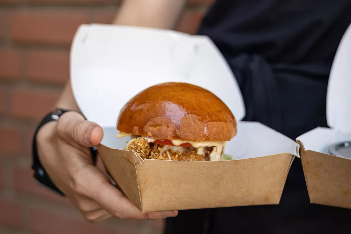Close das mãos de um funcionário segurando uma embalagem de papel kraft aberta com um hambúrguer artesanal suculento, representando a produção de uma marca em uma dark kitchen.