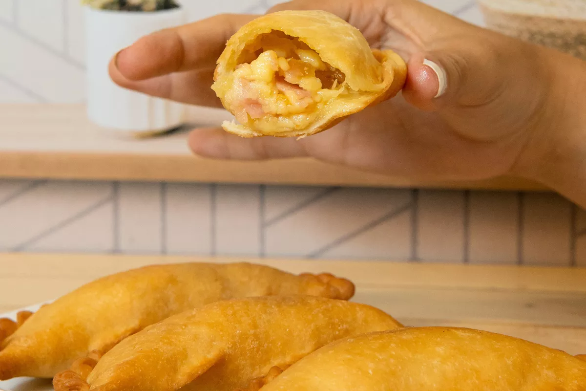 close de uma mão segurando uma empanada frita aberta, revelando o recheio de queijo e presunto derretido, exemplificando o foco em um produto "herói" no delivery hiperespecializado.
