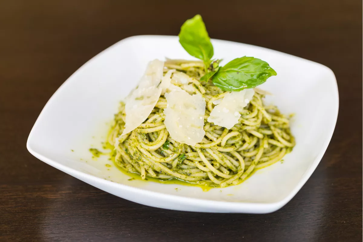 Prato branco quadrado servindo uma porção de macarrão tipo linguine envolvido em molho pesto verde, finalizado com lascas de queijo parmesão e folhas frescas de manjericão. Ótima opção de prato vegetariano para o cardápio de restaurantes.