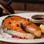 Posta de salmão grelhado com aspecto suculento, servida em um prato branco redondo, acompanhada de fatias de limão siciliano, um pequeno pote de molho escuro e sementes de romã. Exemplo de prato com peixe nobre para atrair clientes na Quaresma.