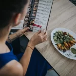 Cliente lendo cardápio impresso ao lado de prato servido, ilustrando o momento de escolha e a importância de um cardápio variado e bem apresentado
