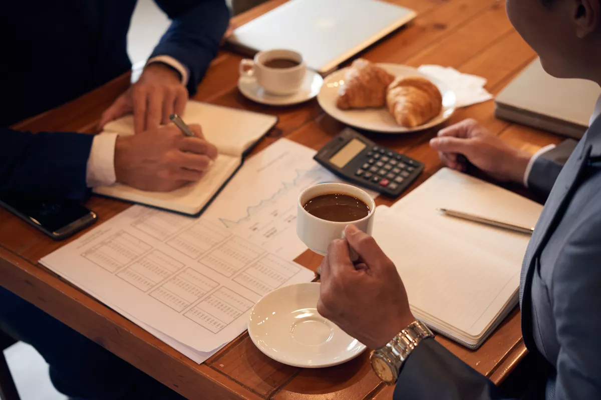 Dois gestores analisando planilhas de custos sobre mesa de madeira em ambiente de cafeteria, com calculadora, cadernos de anotações, xícaras de café e croissants, representando o cálculo da margem de contribuição por prato em um negócio de food service