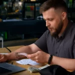 Dono de restaurante com barba e camisa cinza analisando notas fiscais e cupons ao lado de laptop e dinheiro em espécie sobre mesa de madeira, com ambiente de bar desfocado ao fundo, representando o controle financeiro e o acompanhamento da margem de contribuição com auxílio de sistema de gestão