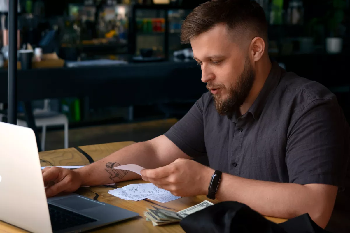 Dono de restaurante com barba e camisa cinza analisando notas fiscais e cupons ao lado de laptop e dinheiro em espécie sobre mesa de madeira, com ambiente de bar desfocado ao fundo, representando o controle financeiro e o acompanhamento da margem de contribuição com auxílio de sistema de gestão