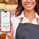 atendente de avental cinza sorrindo e segurando celular com a tela do sistema de comanda eletrônica do ControleNaMão aberta.