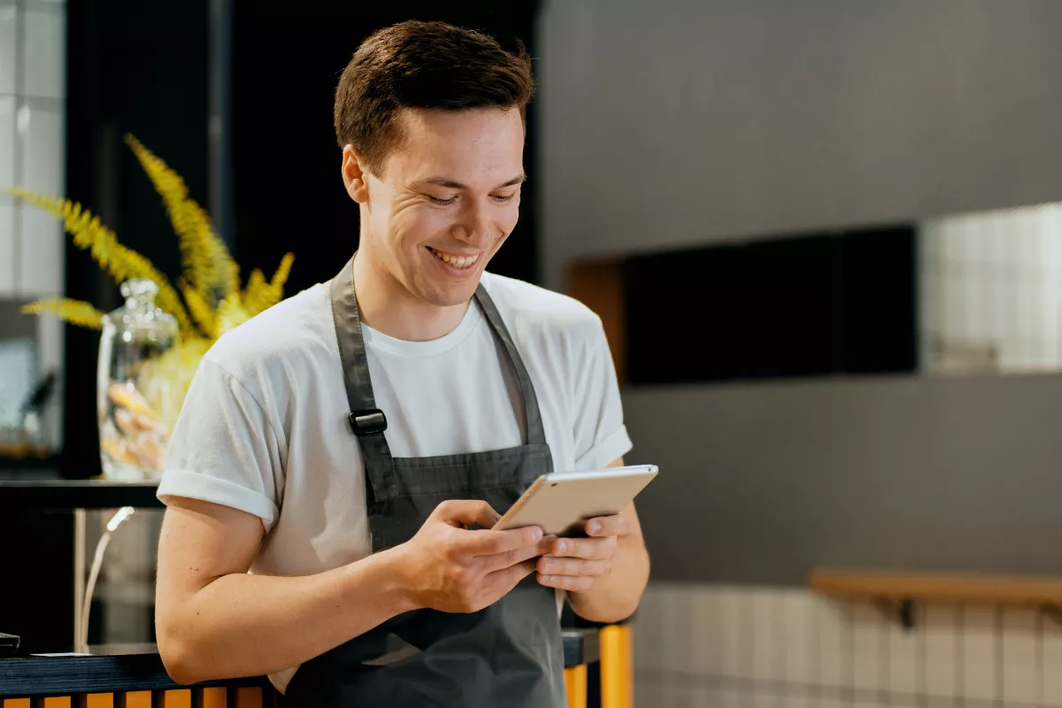 garçom de avental cinza sorrindo enquanto usa tablet no salão do restaurante, com plantas e balcão ao fundo. 