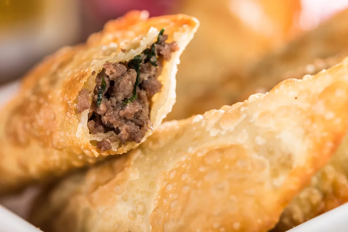 Pastel de carne aberto ao meio mostrando recheio generoso de carne moída com cheiro-verde, com massa frita dourada e crocante, servido em cumbuca branca com fundo desfocado em ambiente de bar ou lanchonete
