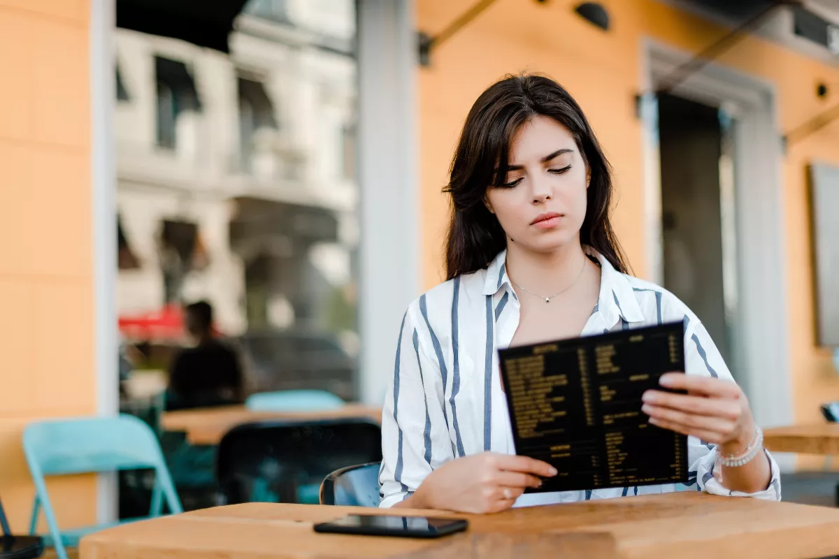 Cliente jovem sentada em mesa de restaurante ao ar livre lendo cardápio extenso com expressão de indecisão, com smartphone sobre a mesa e fachada colorida ao fundo