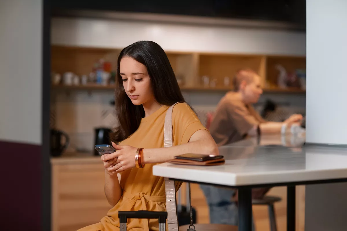 Jovem com bolsa a tiracolo usando smartphone em ambiente de lanchonete ou café, com balcão e outra pessoa ao fundo, representando cliente fazendo pedido por aplicativo de delivery
