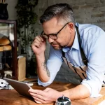 Dono de restaurante com avental listrado e óculos debruçado sobre balcão analisando tablet com expressão concentrada e preocupada, em ambiente de padaria ou café com pães e plantas ao fundo