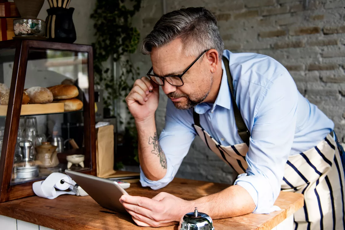 Dono de restaurante com avental listrado e óculos debruçado sobre balcão analisando tablet com expressão concentrada e preocupada, em ambiente de padaria ou café com pães e plantas ao fundo