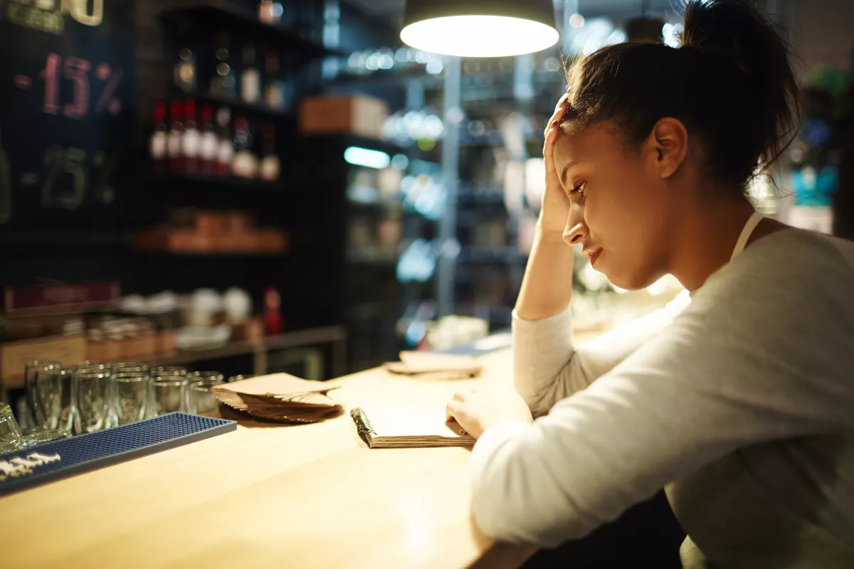 Mulher com avental apoiada no balcão de bar com a cabeça entre as mãos e expressão de esgotamento, com copos e prateleiras de bebidas desfocados ao fundo, representando sobrecarga do dono de restaurante