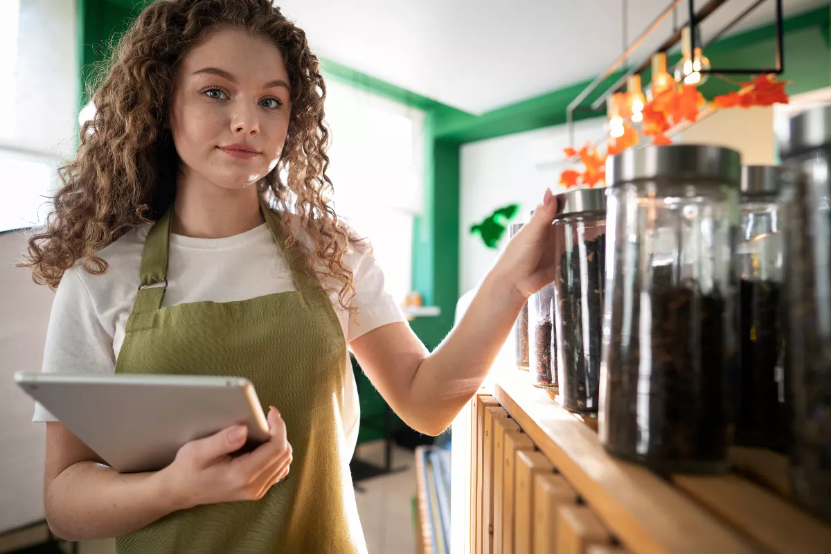 Funcionária de restaurante com avental verde e tablet na mão organizando e verificando produtos em prateleiras de estoque, representando controle de estoque em estabelecimento de food service