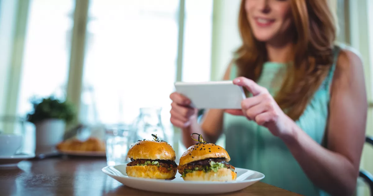 Cliente sorrindo fotografa dois sliders sobre prato branco em mesa de restaurante, segurando o celular na horizontal para registrar o prato antes de comer