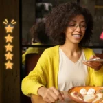 Mulher de óculos e cardigã amarelo sorri enquanto mexe no celular à mesa de restaurante, com prato de comida à frente e ícones de estrelas e medidor de satisfação sobrepostos à imagem
