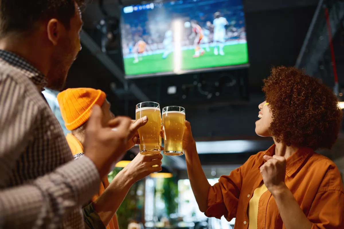 Grupo de amigos brindando com copos de cerveja em bar, com jogo de futebol transmitido em telão ao fundo, em clima de torcida durante Copa do Mundo
