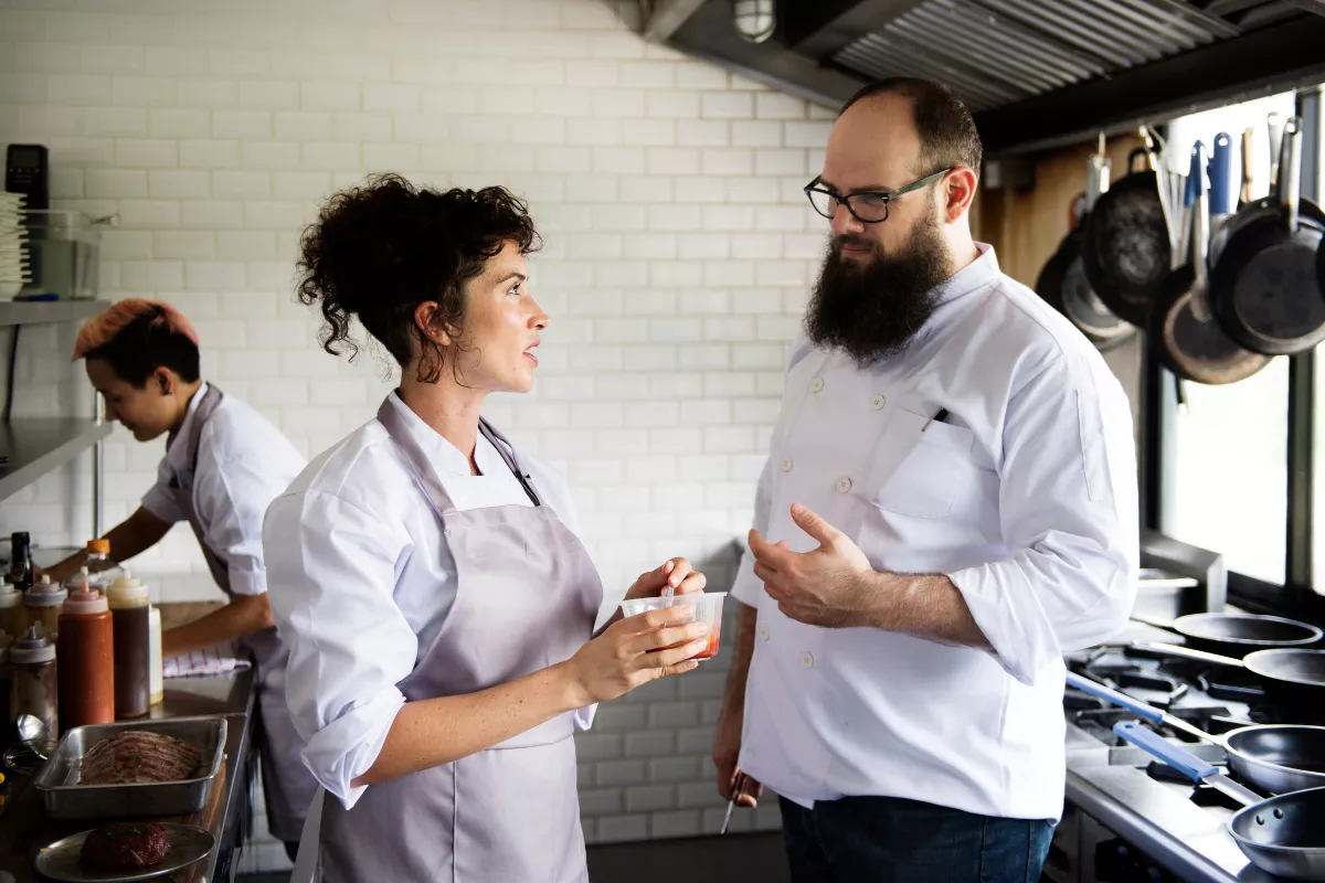 Chef com avental conversando com colega de uniforme branco na cozinha profissional do restaurante, com terceiro funcionário trabalhando ao fundo, representando feedback entre equipe durante o treinamento
