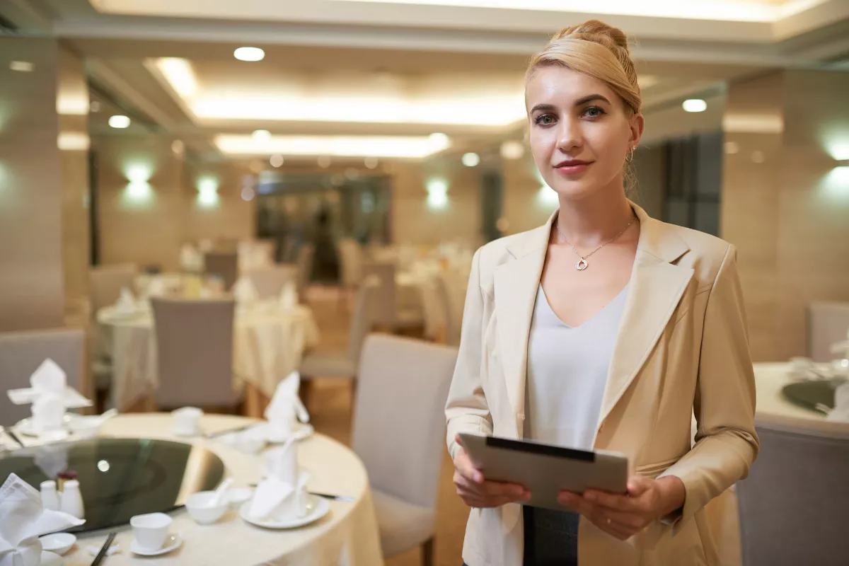 Gerente de restaurante com blazer bege segurando tablet, de pé no salão com mesas postas e cadeiras ao fundo, em ambiente iluminado de restaurante fino