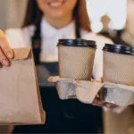 Atendente com avental preto e camisa branca entrega saco kraft e suporte com dois copos térmicos com tampa, no balcão de uma cafeteria com fundo desfocado, ilustrando a entrega de pedido take away