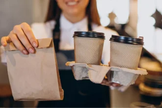 Atendente com avental preto e camisa branca entrega saco kraft e suporte com dois copos térmicos com tampa, no balcão de uma cafeteria com fundo desfocado, ilustrando a entrega de pedido take away