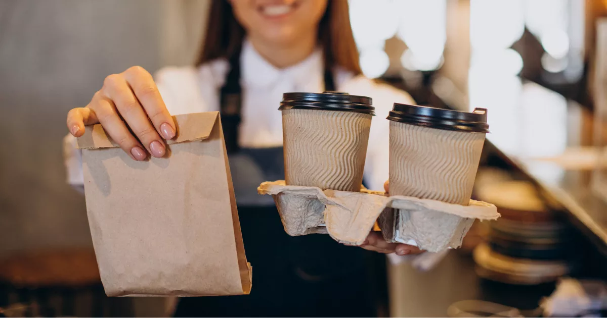 Atendente com avental preto e camisa branca entrega saco kraft e suporte com dois copos térmicos com tampa, no balcão de uma cafeteria com fundo desfocado, ilustrando a entrega de pedido take away