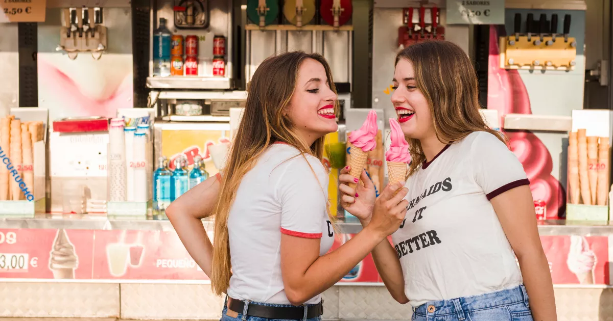 Duas jovens sorrindo segurando casquinhas de sorvete rosa em frente ao balcão de uma sorveteria com máquinas de sorvete expresso e bebidas ao fundo