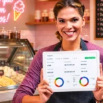 Dona de sorveteria de avental sorrindo segurando tablet com dashboard de analytics, com balcão refrigerado de sorvetes coloridos e cardápio em lousa ao fundo
