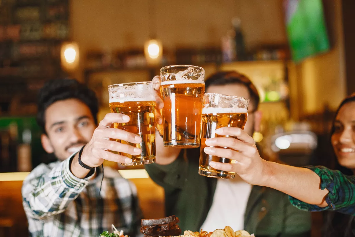 Três amigos sorrindo fazem um brinde com canecas de cerveja clara em um bar iluminado, com petiscos na mesa ao fundo
