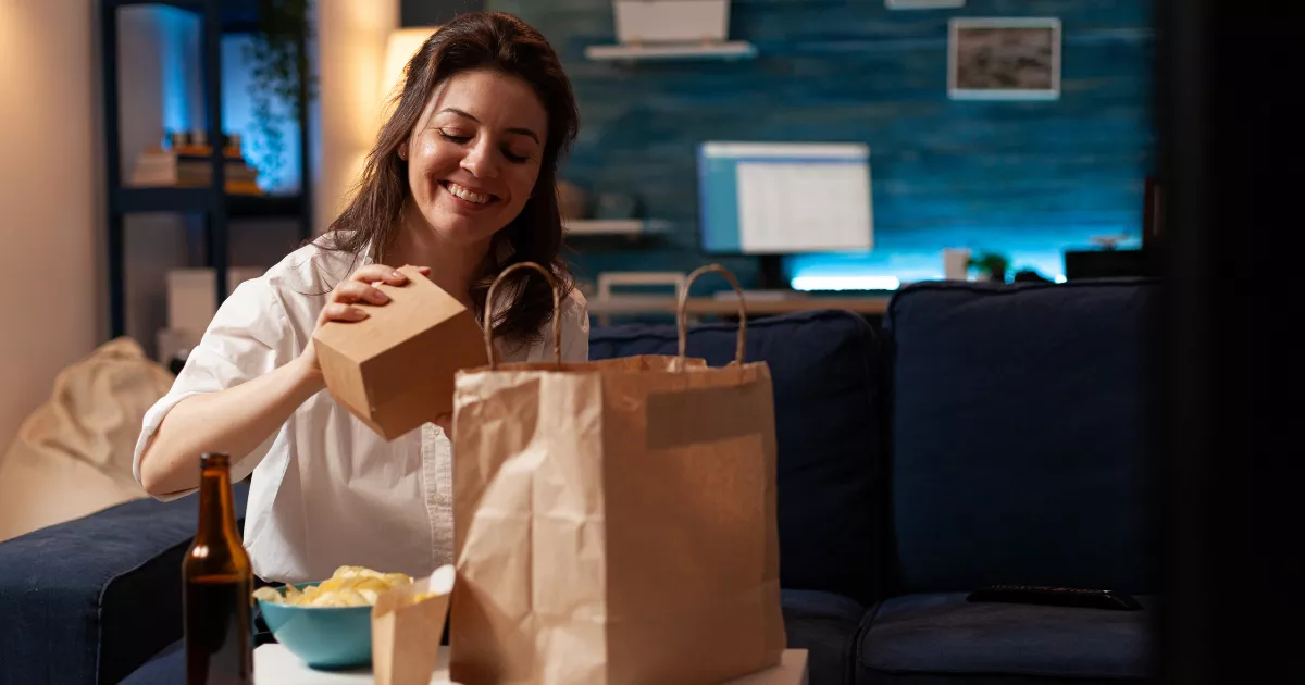 Mulher de camisa branca sentada no sofá da sala, sorrindo ao abrir caixa de papelão kraft retirada de sacola de papel, com garrafa de cerveja e petiscos sobre a mesa de centro, ilustrando consumo de pedido take away em casa