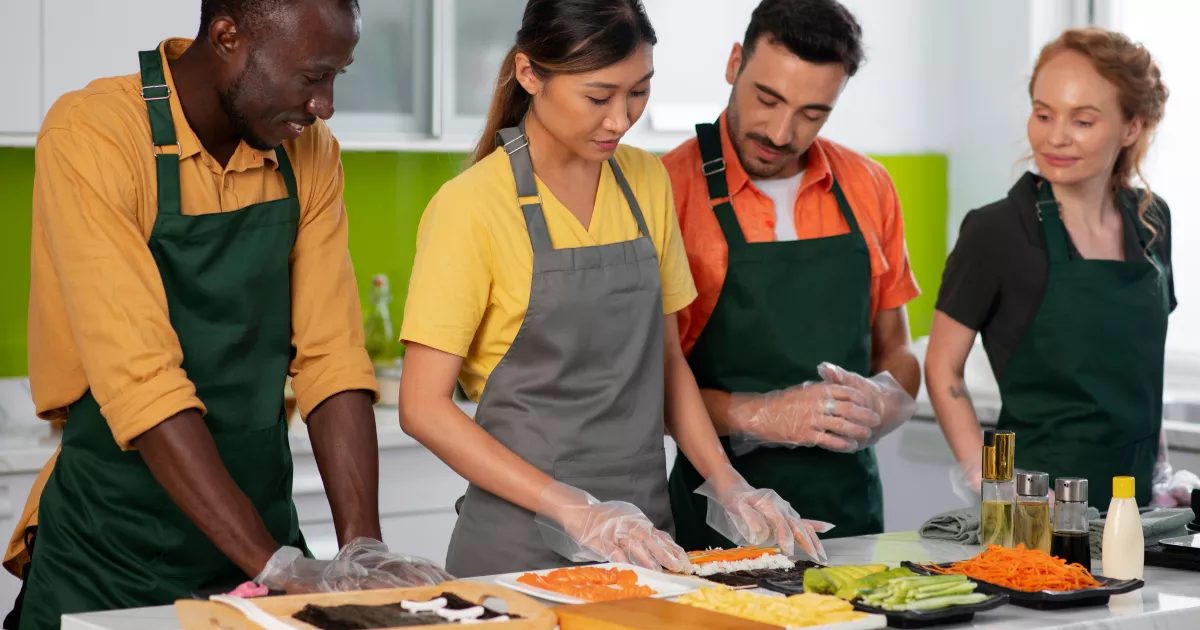 Quatro funcionários de restaurante com aventais usando luvas plásticas descartáveis para preparar rolinhos de sushi sobre bancada branca, com ingredientes frescos dispostos à frente