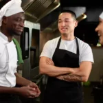 Três cozinheiros uniformizados conversando descontraidamente na cozinha do restaurante, com equipamentos industriais ao fundo, representando integração de funcionário novo à equipe