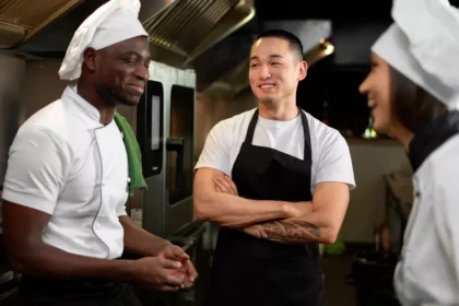 Três cozinheiros uniformizados conversando descontraidamente na cozinha do restaurante, com equipamentos industriais ao fundo, representando integração de funcionário novo à equipe