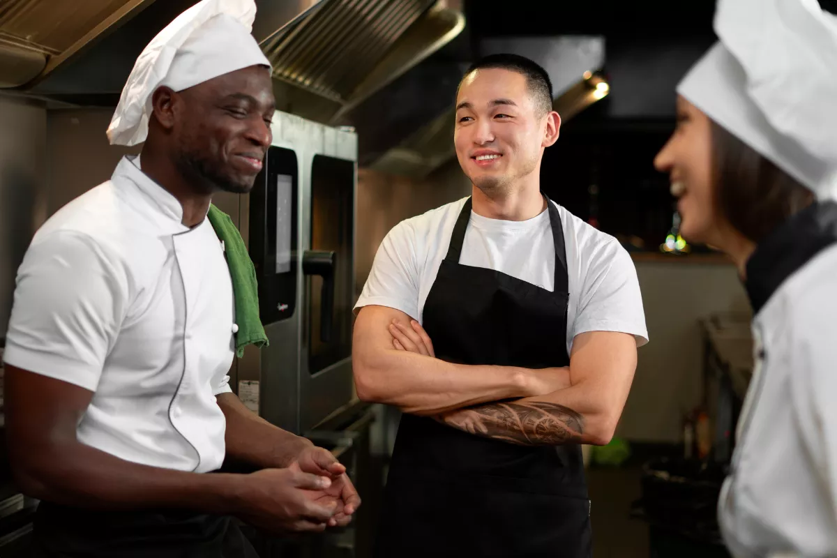Três cozinheiros uniformizados conversando descontraidamente na cozinha do restaurante, com equipamentos industriais ao fundo, representando integração de funcionário novo à equipe