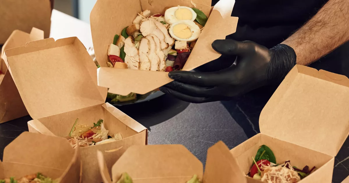 Cozinheiro com luva preta encaixando salada de frango grelhado, ovo cozido e folhas verdes em caixa kraft aberta, com outras embalagens similares montadas ao redor sobre bancada escura, ilustrando o processo de embalagem de pedidos take away na cozinha