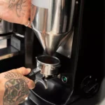 Mãos tatuadas de barista posicionando portafiltro no dosador de moedor de café profissional preto, com segundo moedor ao fundo em ambiente de cafeteria