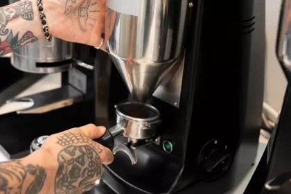 Mãos tatuadas de barista posicionando portafiltro no dosador de moedor de café profissional preto, com segundo moedor ao fundo em ambiente de cafeteria