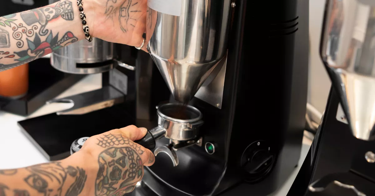 Mãos tatuadas de barista posicionando portafiltro no dosador de moedor de café profissional preto, com segundo moedor ao fundo em ambiente de cafeteria