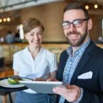 Gerente de restaurante de óculos e blazer escuro segurando tablet e bandeja com prato, ao lado de garçonete com uniforme branco sorrindo no salão do estabelecimento