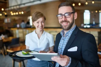 Gerente de restaurante de óculos e blazer escuro segurando tablet e bandeja com prato, ao lado de garçonete com uniforme branco sorrindo no salão do estabelecimento