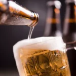 Cerveja clara sendo servida de uma garrafa em uma caneca de vidro com espuma abundante, com outras garrafas desfocadas ao fundo em fundo escuro