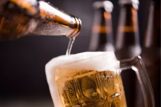 Cerveja clara sendo servida de uma garrafa em uma caneca de vidro com espuma abundante, com outras garrafas desfocadas ao fundo em fundo escuro