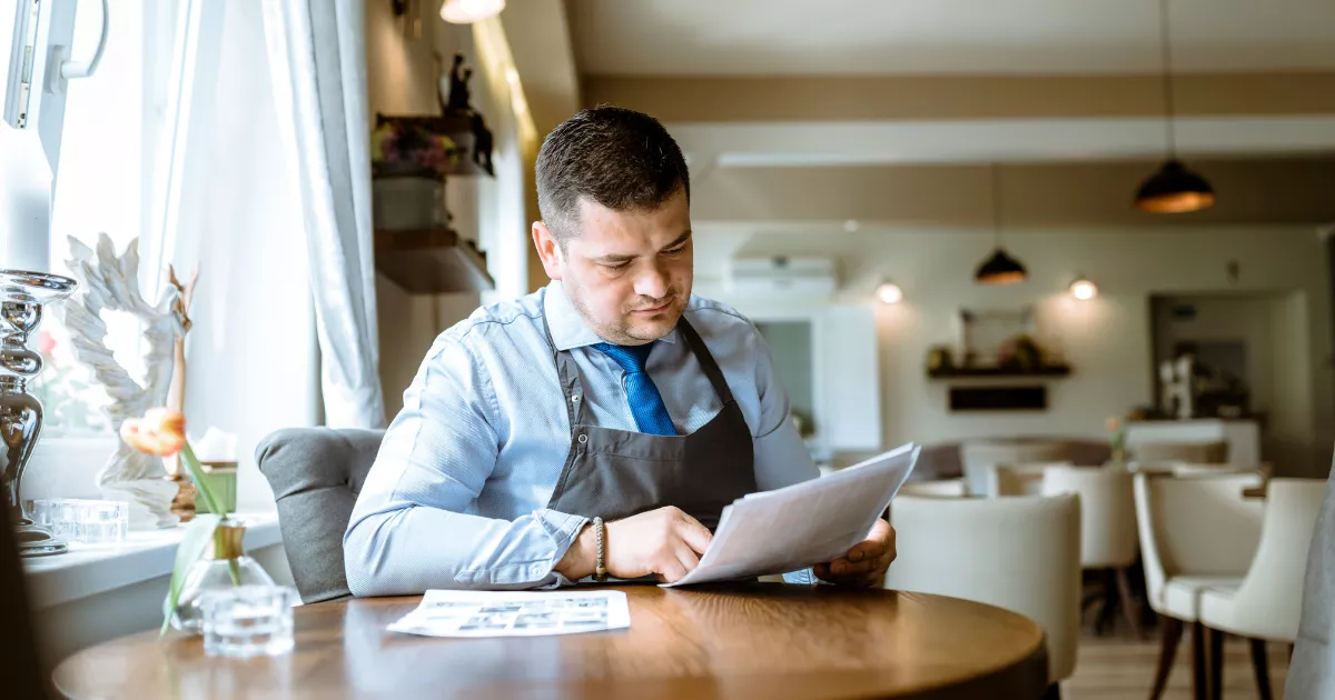 Dono de restaurante com avental, camisa social e gravata azul sentado à mesa redonda de madeira analisando documentos impressos, com mesas e cadeiras brancas do salão ao fundo