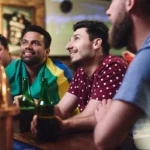 Torcedores sorridentes sentados no balcão de bar assistindo jogo de futebol, um deles com bandeira do Brasil nos ombros e garrafas de cerveja verde na mão