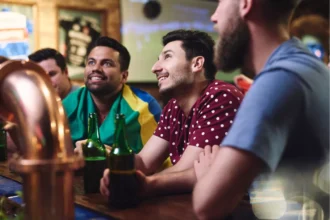 Torcedores sorridentes sentados no balcão de bar assistindo jogo de futebol, um deles com bandeira do Brasil nos ombros e garrafas de cerveja verde na mão